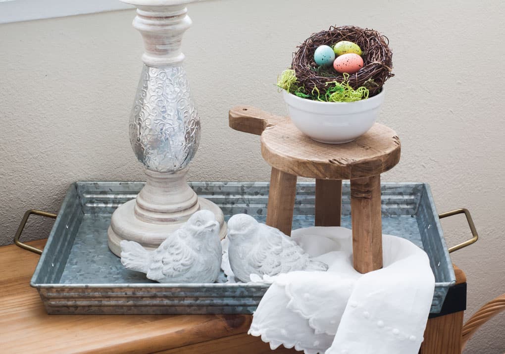 A galvanized tray with handles holding a candle holder miniature stool nest bowl and cement birds on a wood bench