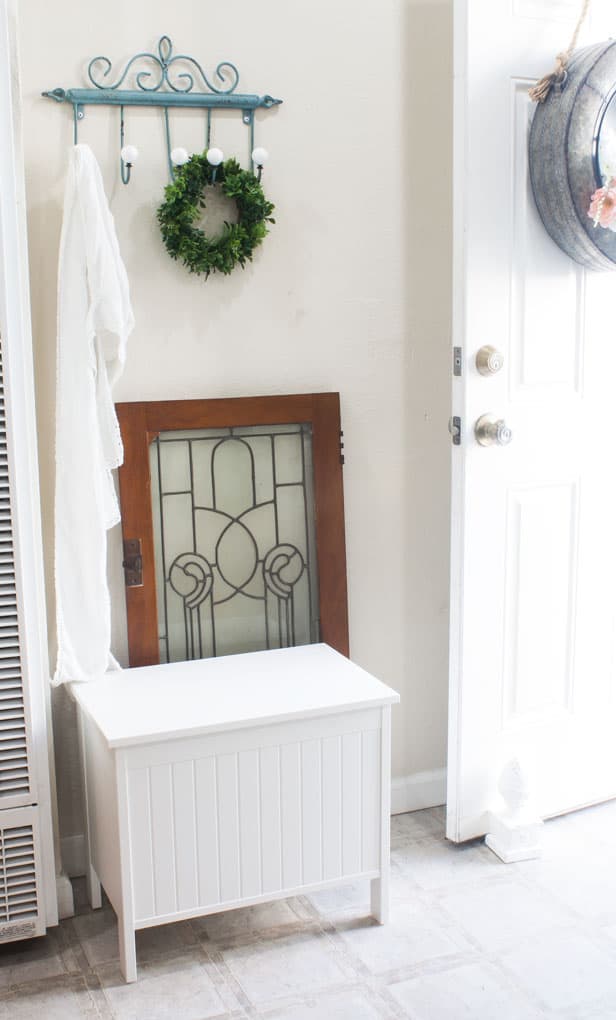 An entry way door opens to a mudroom nook with a seating bench lead glass window hooks and decorative items