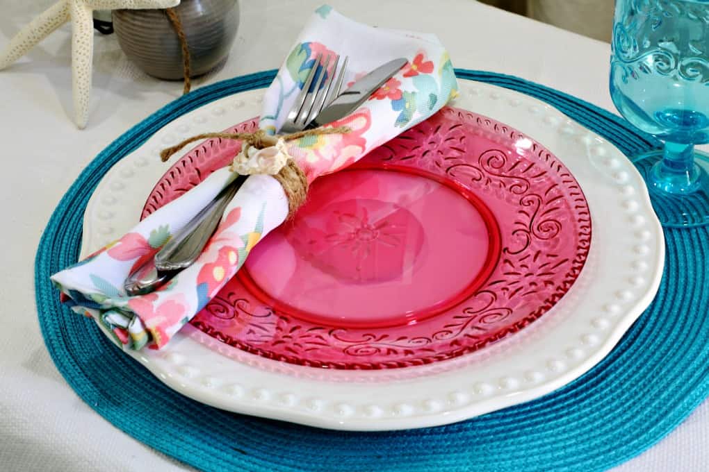 tablesetting on table with white tablecloth teal placemat pink and white dish napkin goblet with flower vase and starfish