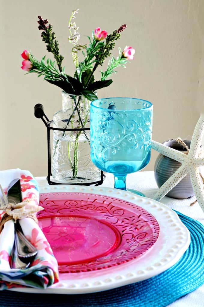 tablesetting on table with white tablecloth teal placemat pink and white dish napkin goblet with flower vase and starfish