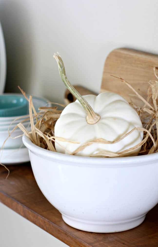 adding fall decor to a small kitchen pumpkin in white bowl with rafia sitting on wood surface with cutting board and dinnerware