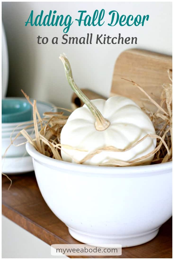 adding fall decor to a small kitchen pumpkin in white bowl with rafia sitting on wood surface with cutting board and dinnerware