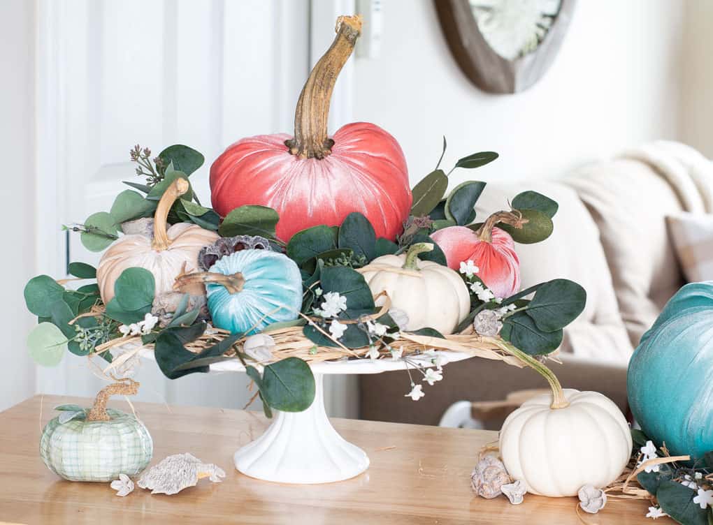 diy velvet pumpkins just like the pros velvet pumpkins in coral aqua and natural on a white cake stand with leaves flowers and natural elements on top of an wood surface with sofa in background