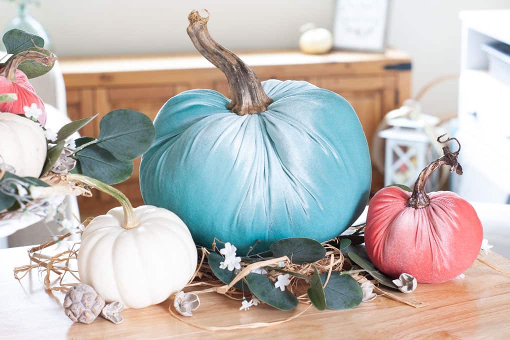 diy velvet pumpkins just like the pros velvet pumpkins in coral aqua and natural on a white cake stand with leaves flowers and natural elements on top of an wood surface with sofa in background