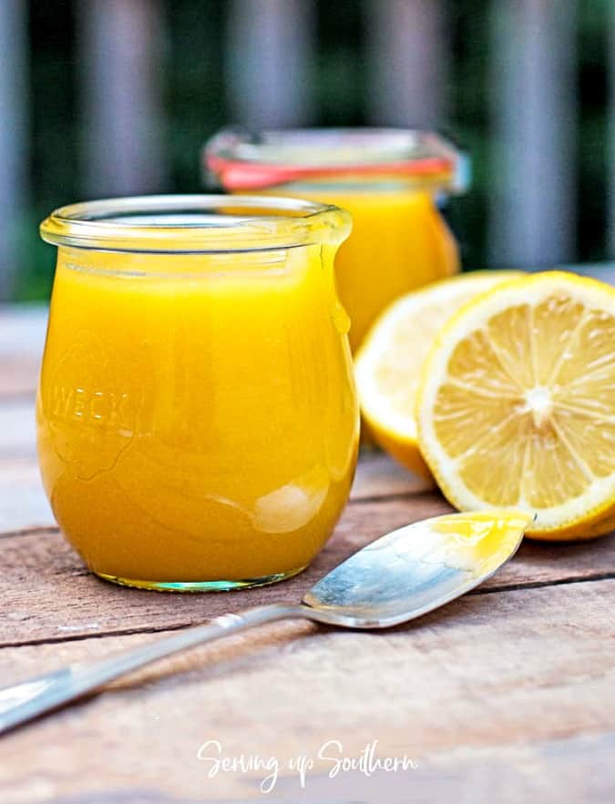 Open jar of lemon curd and a spoonful of curd sitting on a wood board with lemons in the background.