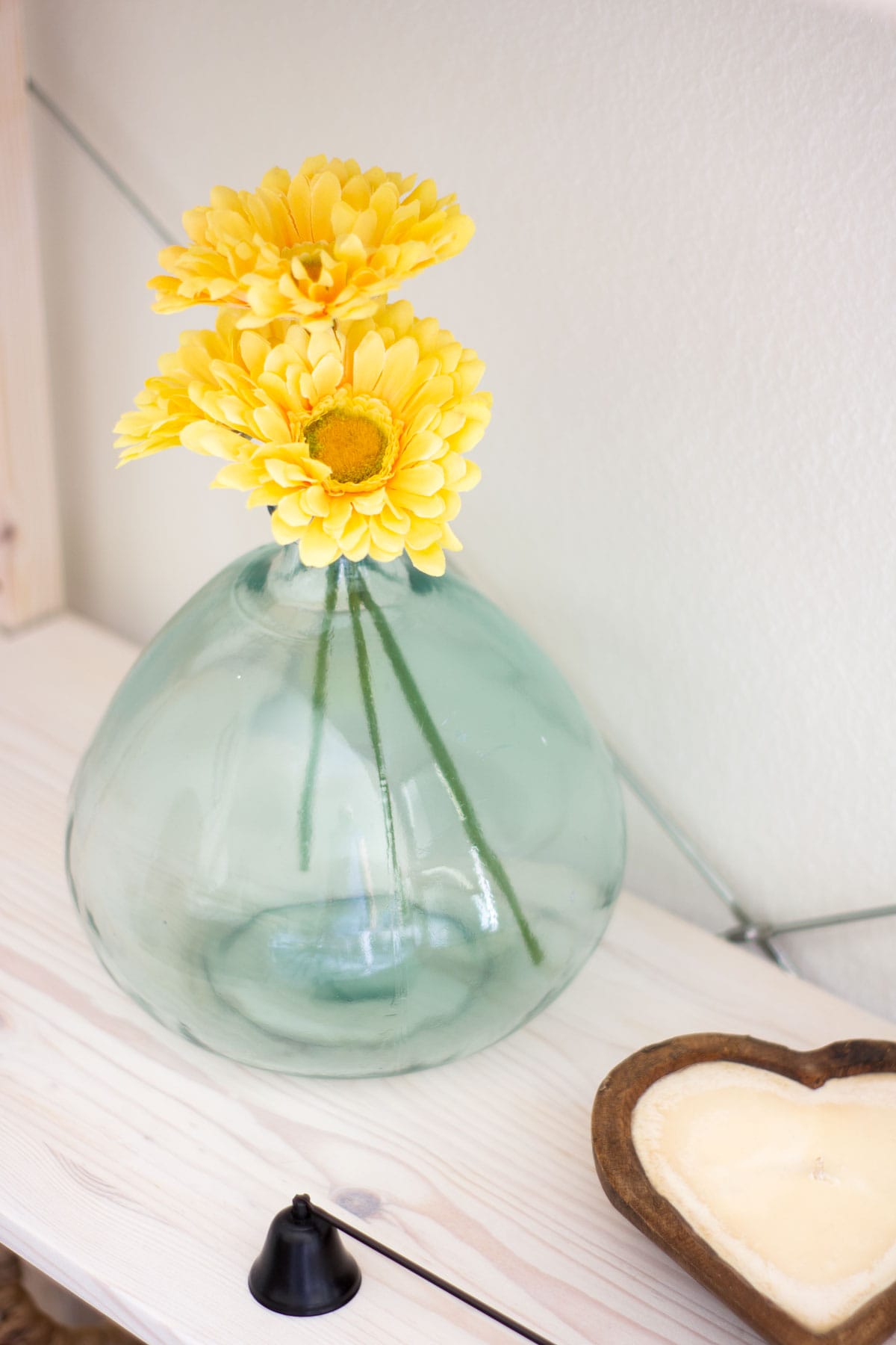 glass vase with yellow daisies and heart shaped candle on wood shelf