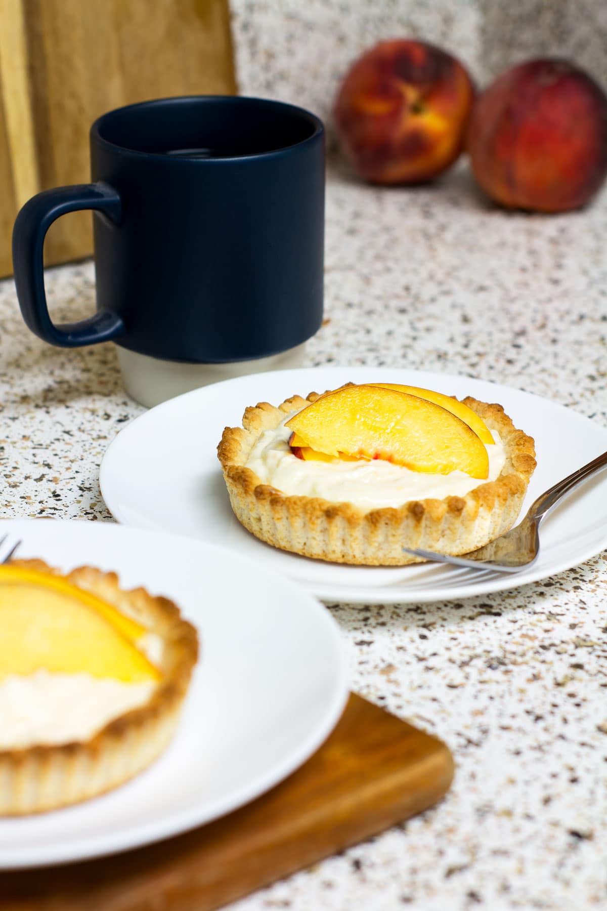 fresh peach mini tart on white plate with fork and coffee cup and peaches in background