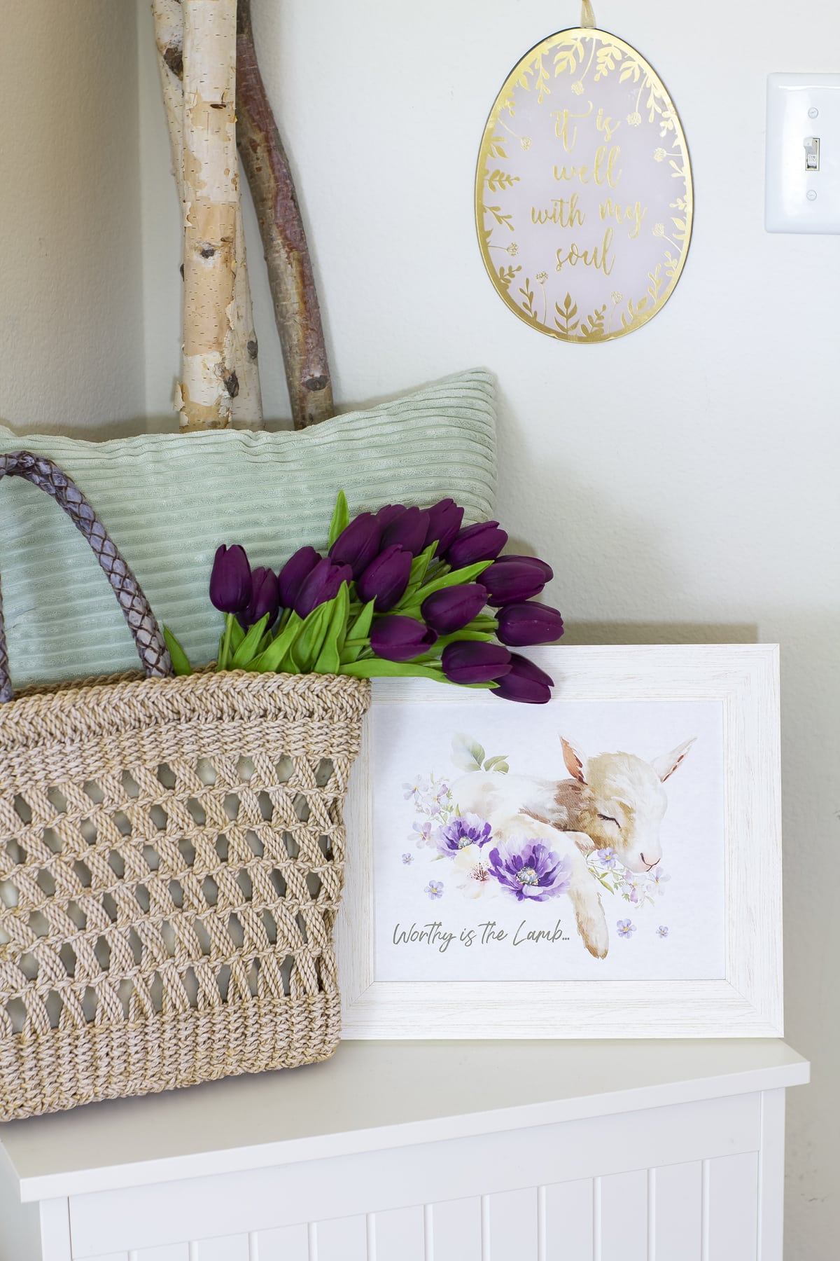 white bench with woven tote and purple tulips with tiny lamb artwork in front of mint pillow