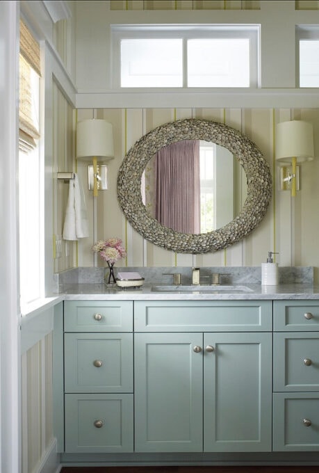 bathroom with sage green cabinets and large oval vintage mirror with striped wallpaper and shaded sconces
